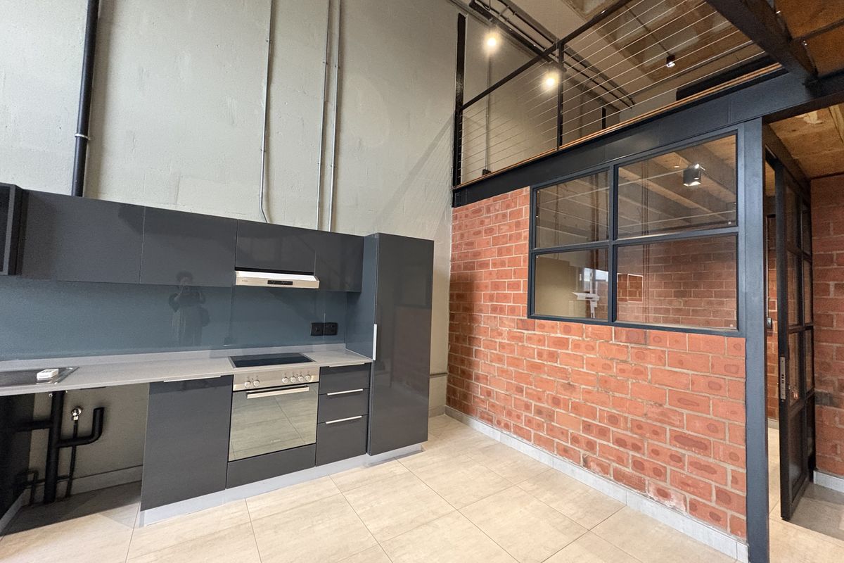 Modern industrial-style kitchen featuring high-gloss cabinetry, built-in oven and hob, exposed brick wall, and a mezzanine level with steel railings.