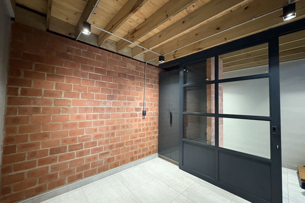 Modern interior space featuring an exposed brick wall, industrial-style timber ceiling beams, tiled flooring, and a sleek black-framed glass partition door.