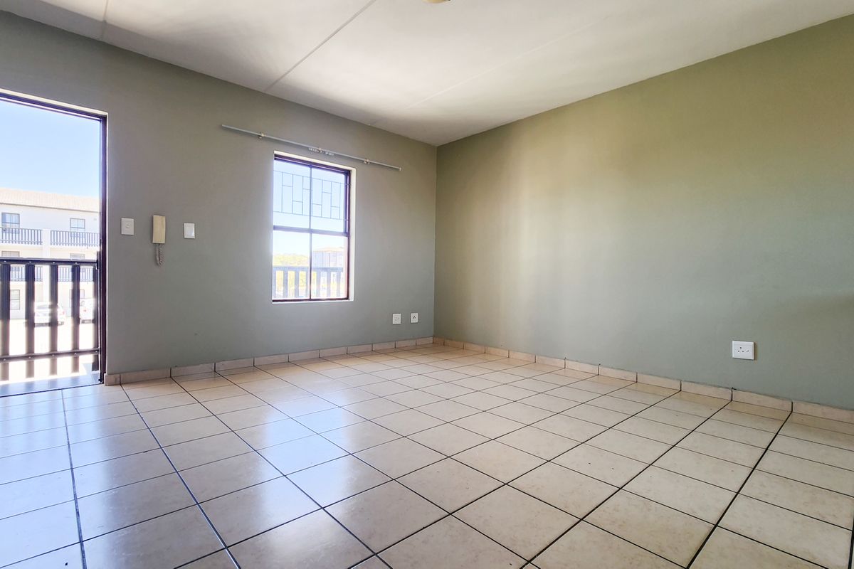 An empty, open-plan living area featuring neutral painted walls, tiled flooring, and direct access to a balcony through a security gate.