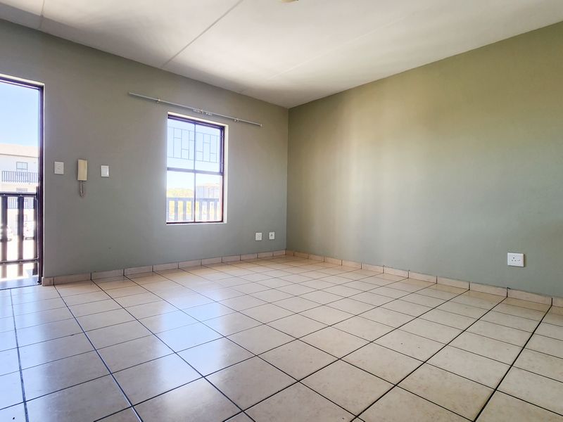 An empty, open-plan living area featuring neutral painted walls, tiled flooring, and direct access to a balcony through a security gate.