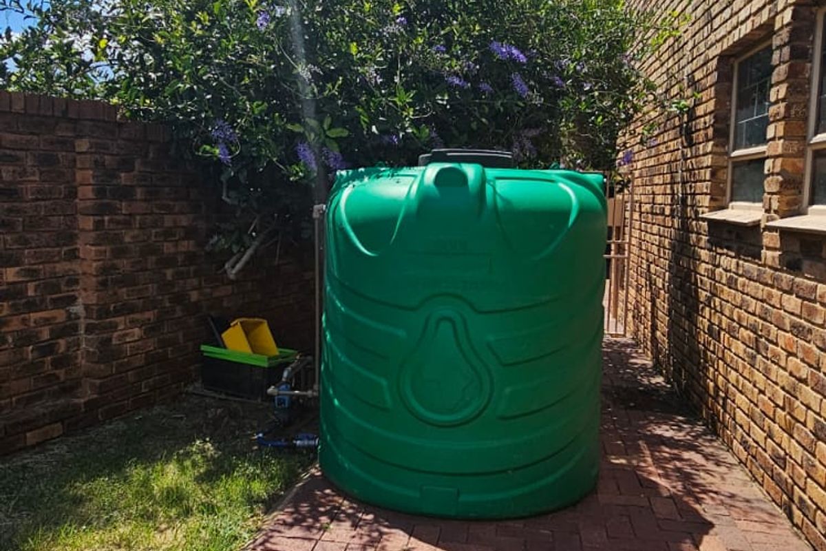 Exterior side area featuring a green water storage tank installed on a paved walkway alongside the brick house wall.