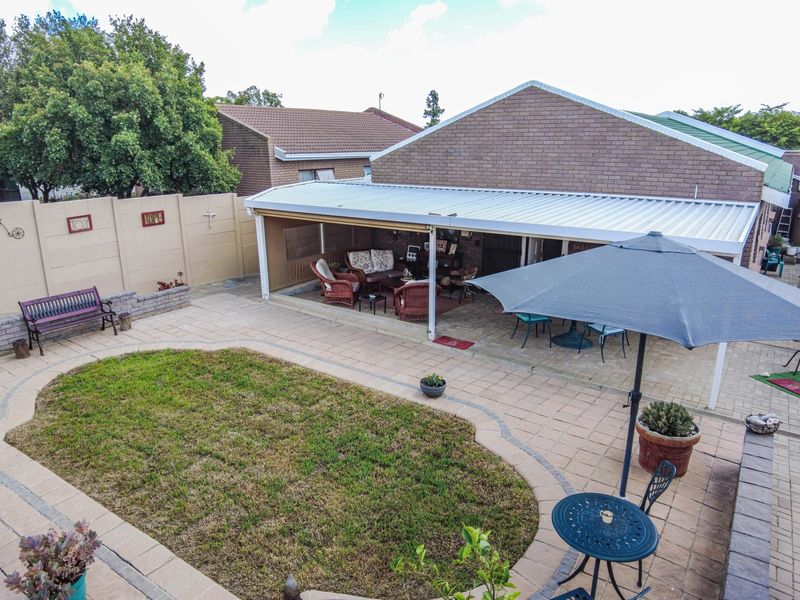 A rear view of the property featuring a paved courtyard, a covered patio area, and a landscaped garden with a boundary wall.
