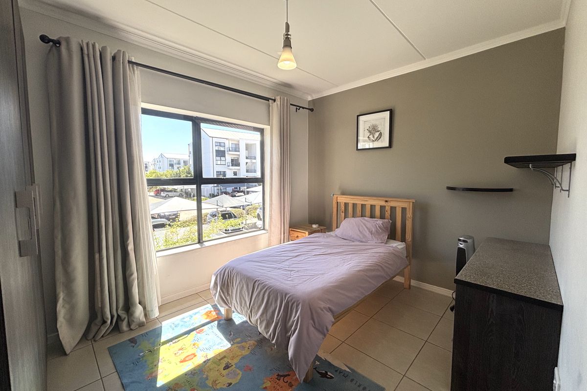 A well-lit bedroom featuring tiled flooring, neutral wall tones, and a large window providing natural light and views of the surrounding complex.