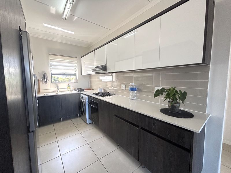Modern galley-style kitchen featuring sleek dark wood lower cabinets, white upper cabinetry, stone countertops, gas hob, and integrated laundry appliances.