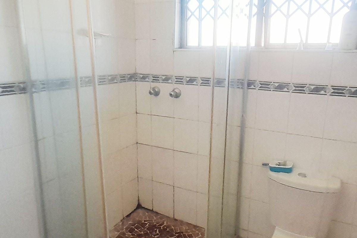 A functional bathroom featuring a corner shower enclosure, tiled walls and flooring, and a standard toilet with security bars on the window.