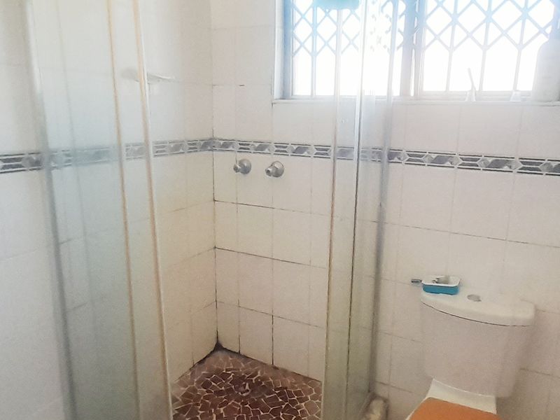 A functional bathroom featuring a corner shower enclosure, tiled walls and flooring, and a standard toilet with security bars on the window.
