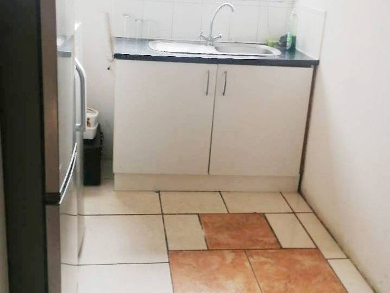 A compact kitchen area featuring basic cabinetry, a sink, and a security gate on the window, with mismatched floor tiling.