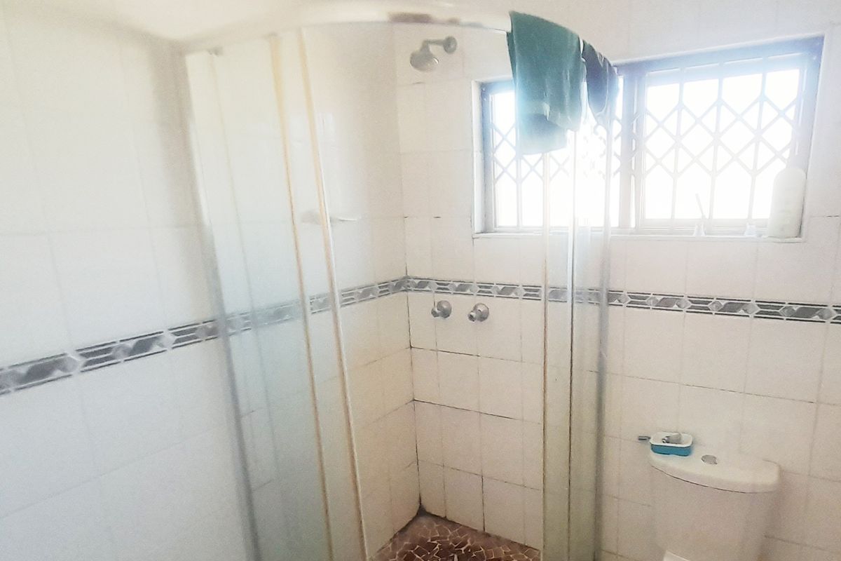 A dated bathroom featuring a corner shower enclosure, toilet, and tiled walls with security bars on the window.
