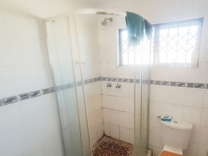 A dated bathroom featuring a corner shower enclosure, toilet, and tiled walls with security bars on the window.