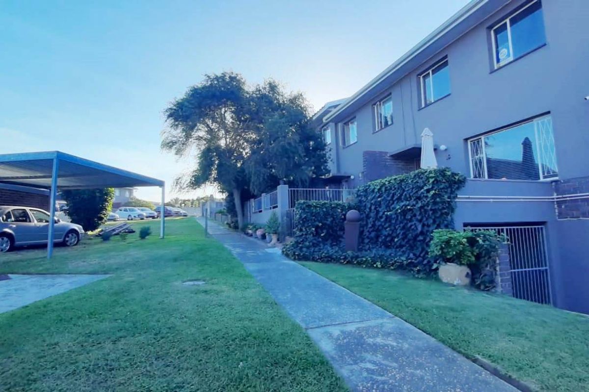 Rear view of the townhouse complex featuring a paved walkway, carport structure, and established garden area with security features.