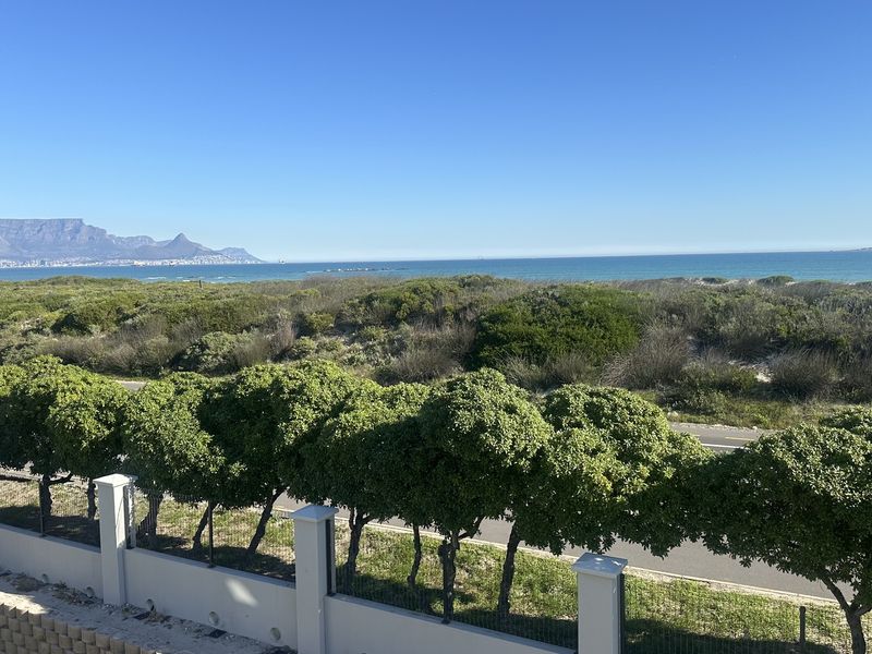 Elevated coastal view overlooking landscaped greenery, a road, and the ocean with Table Mountain in the distance.
