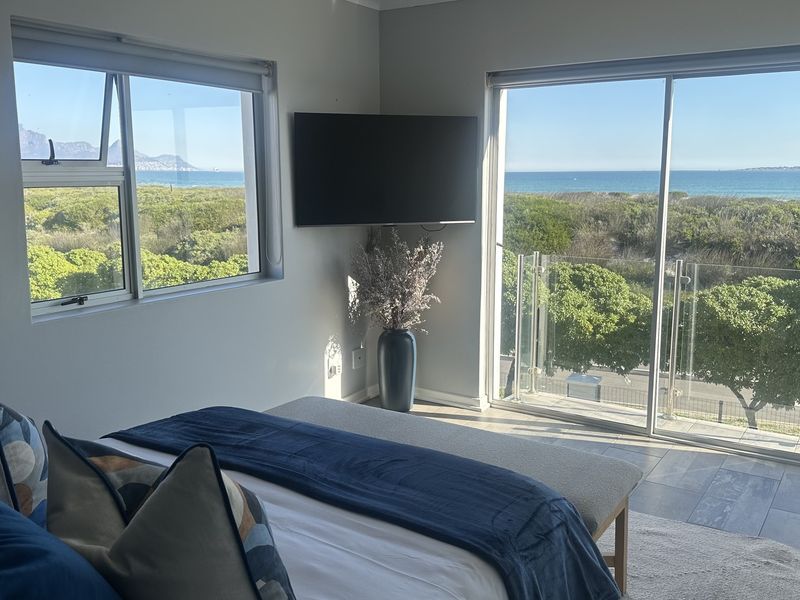 A bright bedroom featuring large sliding glass doors leading to a balcony with a glass balustrade, offering expansive coastal and ocean views.