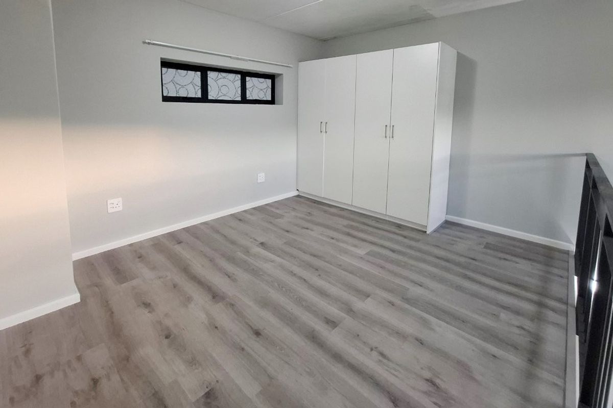 A clean, empty bedroom featuring modern laminate flooring, built-in white cupboards, and a horizontal window, overlooking a lower level via a metal balustrade.