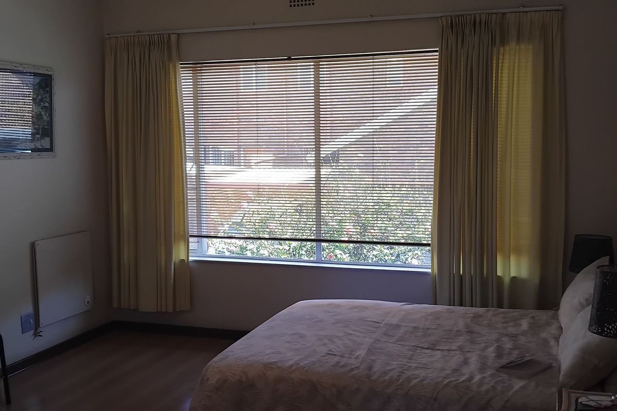 A bedroom featuring laminate flooring, a large window with blinds and curtains, and a wall-mounted heater.