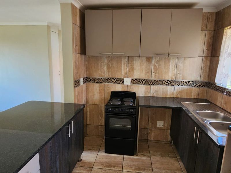 A functional kitchen featuring dark cabinetry, granite countertops, a double basin sink, and a tiled splashback, opening into an adjacent living area.