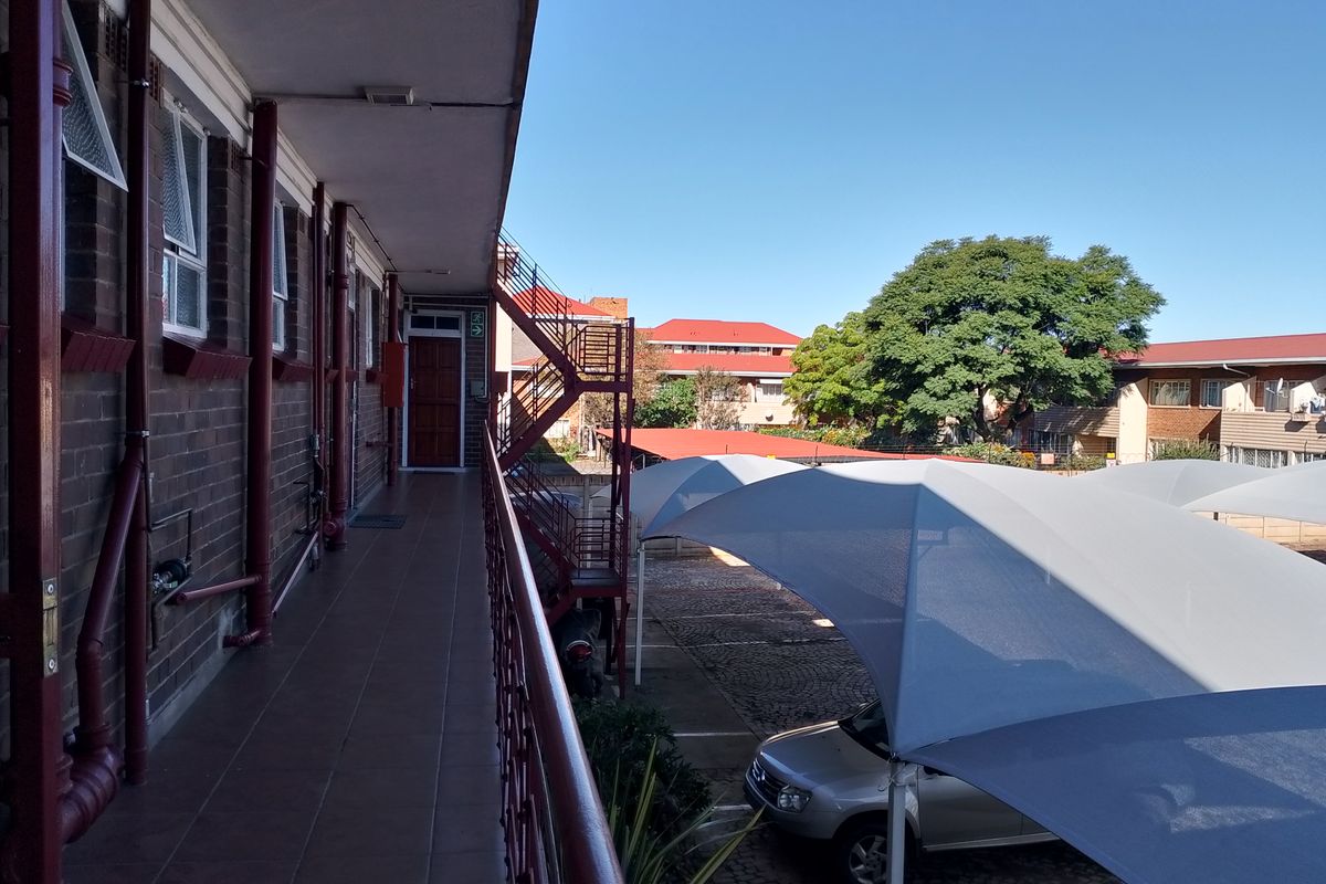 An exterior view of an apartment complex walkway featuring tiled flooring, metal railings, and a view overlooking the parking area with shade sails.