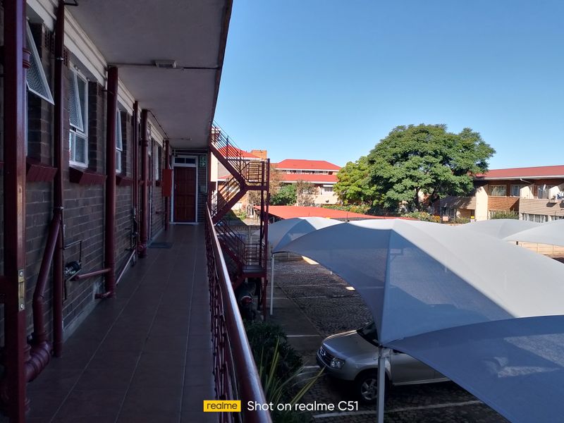 An exterior view of an apartment complex walkway featuring tiled flooring, metal railings, and a view overlooking the parking area with shade sails.