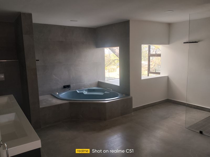 Modern bathroom featuring a corner bathtub, double vanity, and glass shower screen with neutral grey tiling throughout.