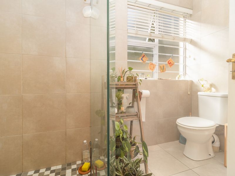 A functional bathroom featuring a glass-enclosed shower, neutral ceramic wall and floor tiling, and a standard toilet with natural light from the window.