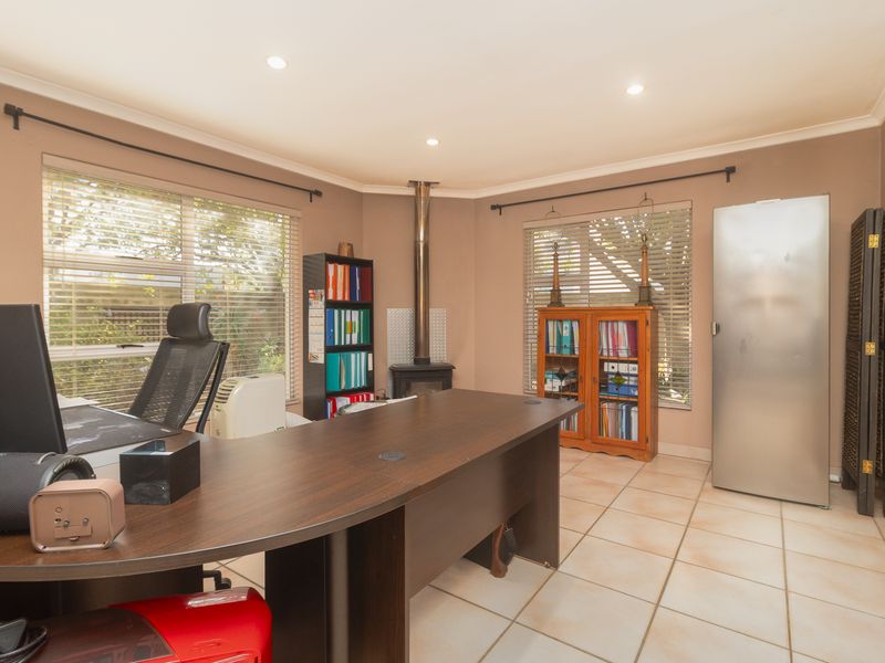 A functional home office space featuring tiled flooring, a central freestanding fireplace, and large windows with venetian blinds providing natural light.
