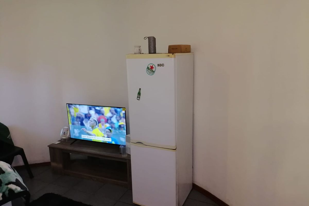 A living area featuring tiled flooring and plain walls, currently housing a refrigerator and television unit.