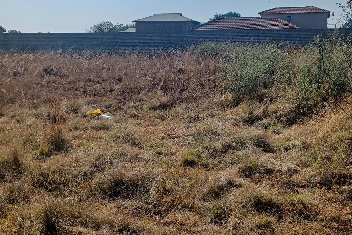 A view of a vacant residential plot featuring dry, overgrown grass and a perimeter boundary wall with neighboring houses visible in the background.