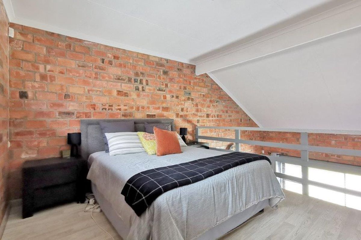A loft-style bedroom featuring an exposed brick feature wall, vaulted ceiling, and a mezzanine-style railing overlooking the lower level.