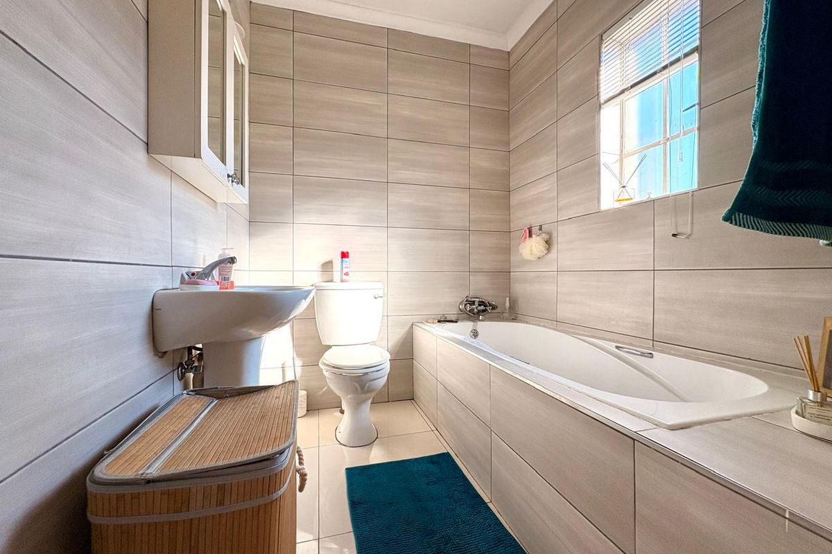 A well-maintained bathroom featuring neutral wall tiles, a built-in bathtub, a pedestal sink, and a toilet, complemented by natural light from a window.