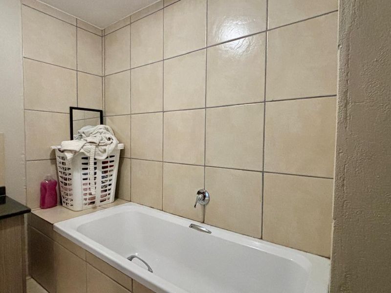 A functional bathroom featuring a built-in bathtub with wall-mounted fixtures, a vanity cabinet with a countertop basin, and neutral ceramic wall tiling.