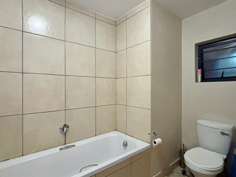 A functional bathroom featuring a bathtub with tiled surround and a standard toilet, complemented by a small window with security bars.