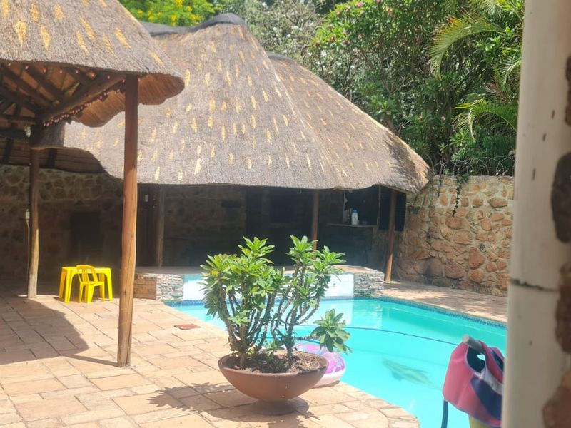 Outdoor entertainment area featuring a swimming pool, paved patio, and a traditional thatched lapa structure set against a stone boundary wall.