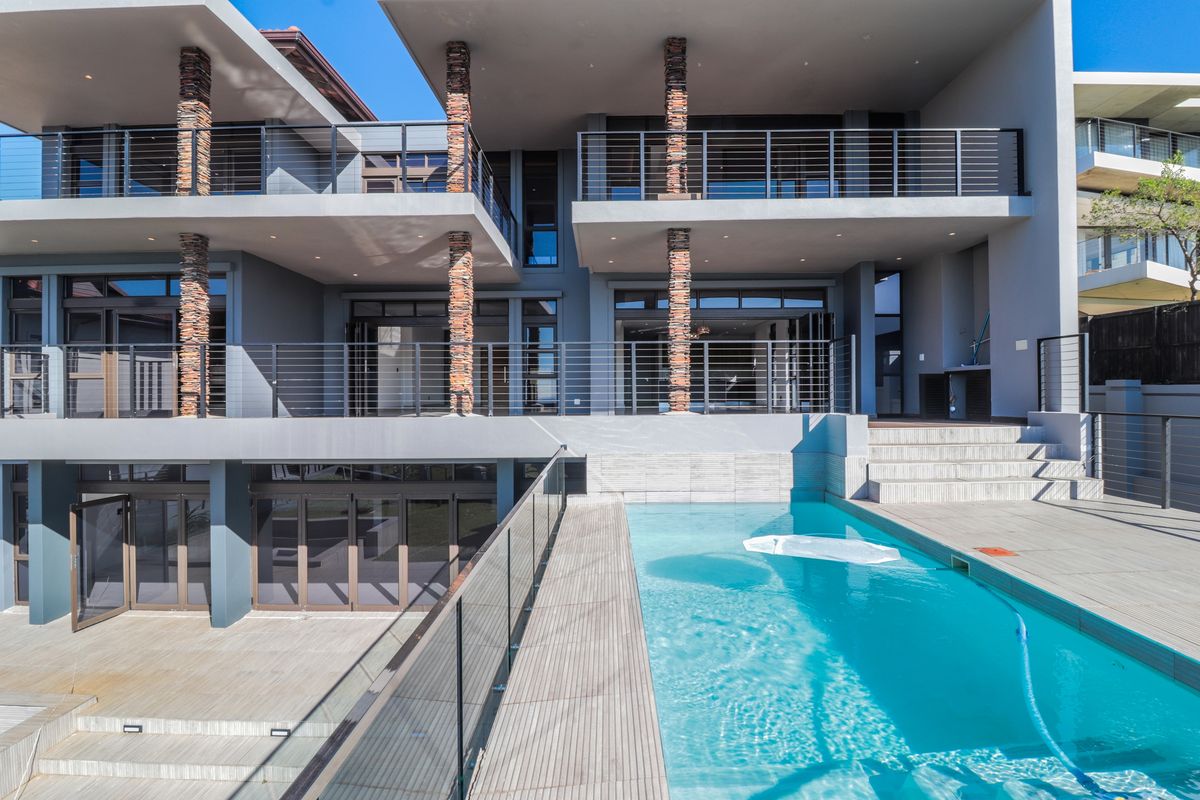 Modern multi-level rear exterior featuring a swimming pool, expansive timber decking, stone-clad columns, and glass balustrades with seamless indoor-outdoor flow.