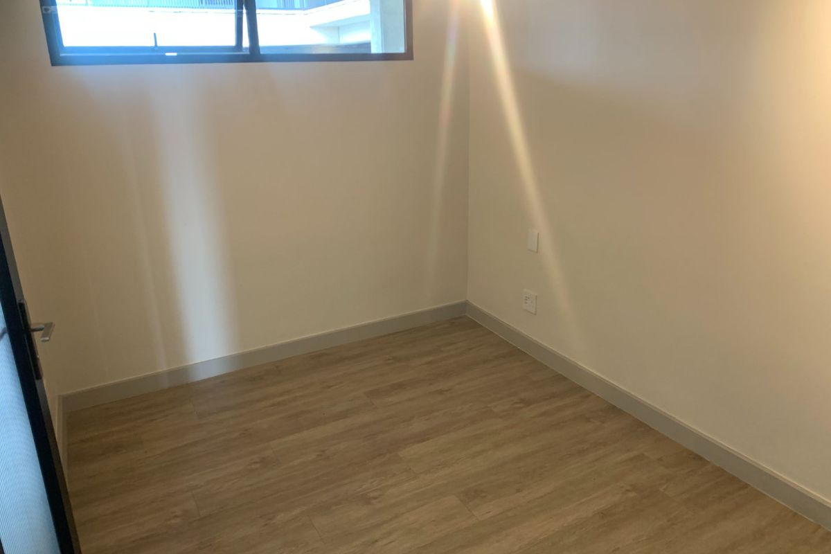 A clean, empty bedroom featuring modern light wood-look flooring, neutral walls, and a high-set horizontal window providing natural light.