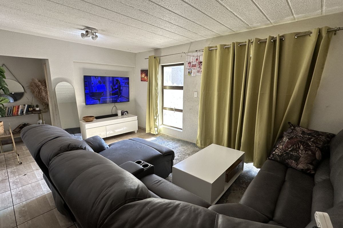 A comfortable living room featuring tiled flooring, a wall-mounted television area, and large windows with floor-to-ceiling curtains providing natural light.