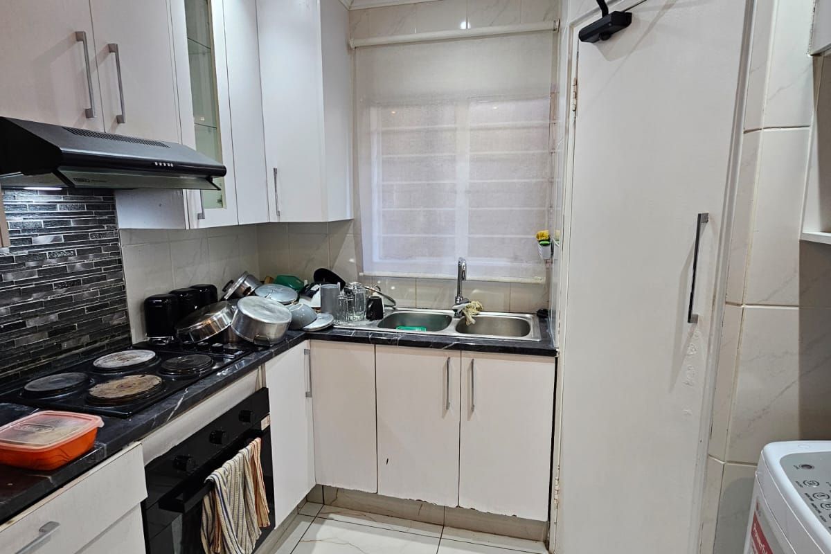 Compact kitchen featuring white cabinetry, dark countertops, tiled backsplash, built-in oven, electric hob, and double basin sink with tiled flooring.