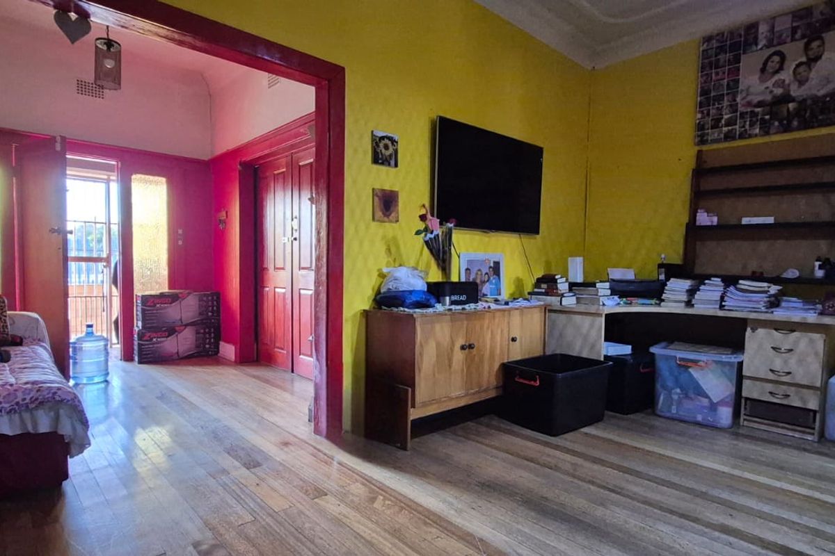 A living area featuring original timber flooring, high ceilings, and a transition into an adjacent room with access to an exterior doorway.