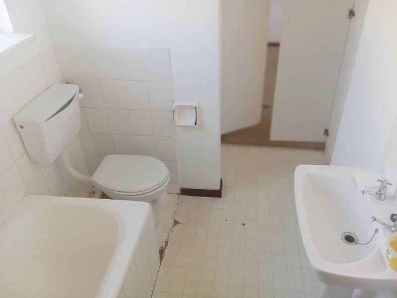A dated bathroom featuring a standard pedestal basin, toilet, and bathtub with white wall tiling, requiring renovation and floor repairs.