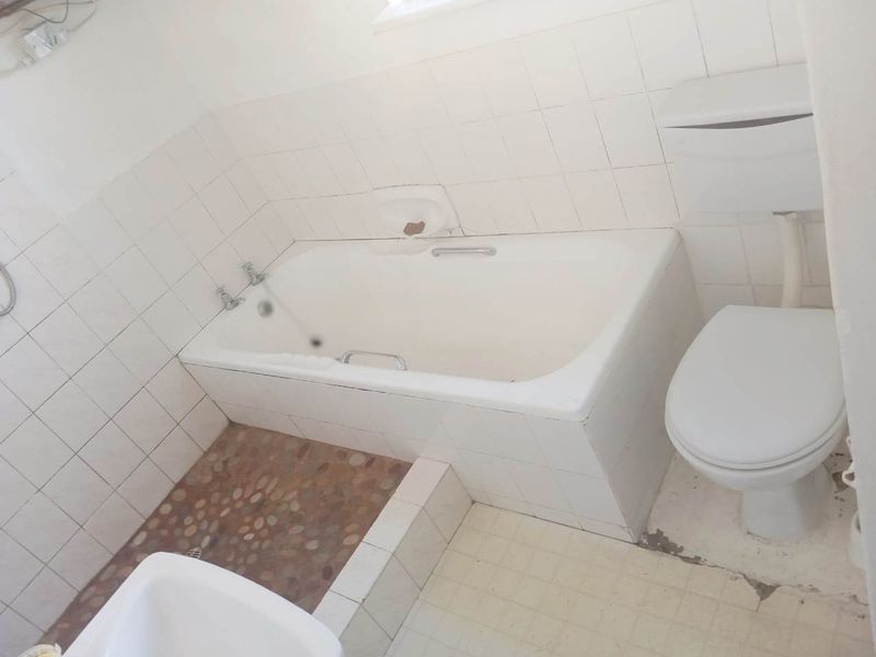 A dated bathroom featuring a built-in bathtub, toilet, and tiled finishes. The space requires renovation to address worn flooring and dated fixtures.
