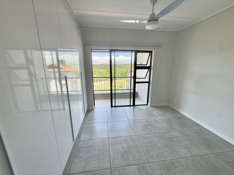 A bright, empty bedroom featuring modern grey tiled flooring, floor-to-ceiling built-in cupboards, a ceiling fan, and a sliding door leading to a balcony.