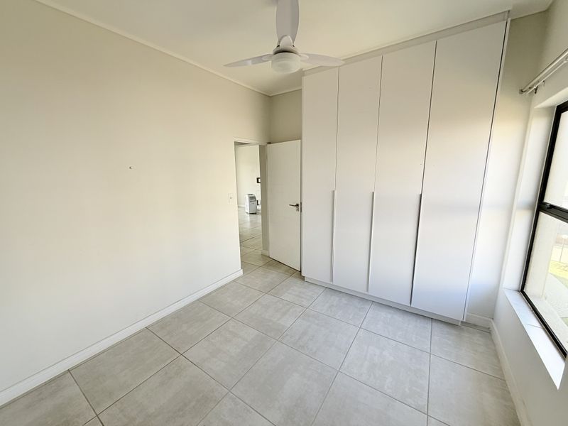 A bright, empty bedroom featuring neutral tiled flooring, built-in white cupboards, and a ceiling fan, offering a clean and versatile living space.