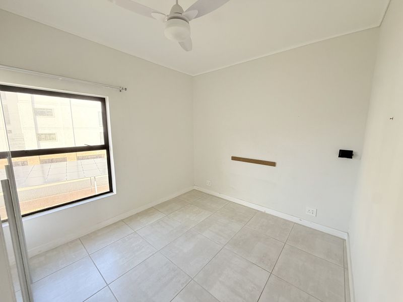 A bright, empty bedroom featuring neutral tiled flooring, a ceiling fan, and large window, providing a clean canvas for personalization.