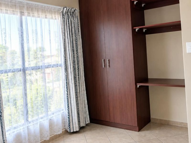 A bright bedroom featuring tiled flooring, a built-in wooden wardrobe with integrated shelving, and large windows allowing for ample natural light.