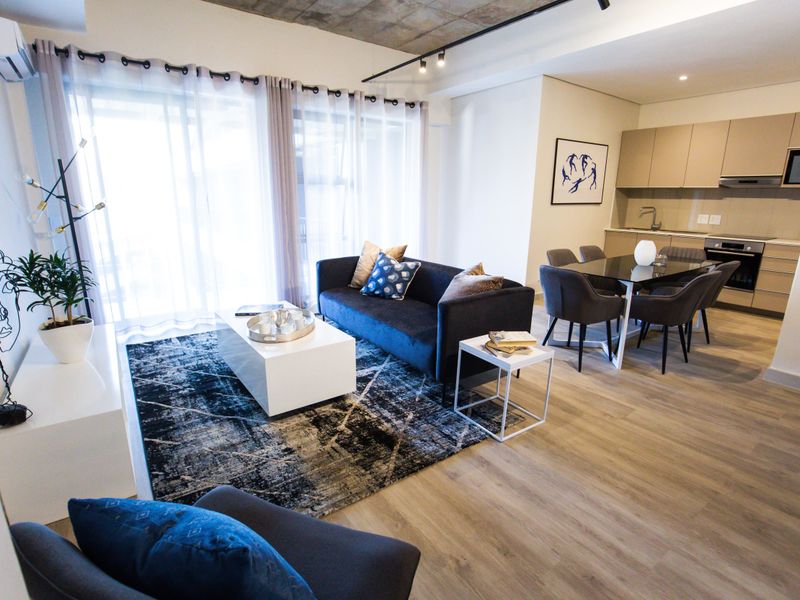 Modern open-plan living area featuring laminate flooring, exposed concrete ceilings, track lighting, and a sleek, fully equipped kitchen with integrated appliances.