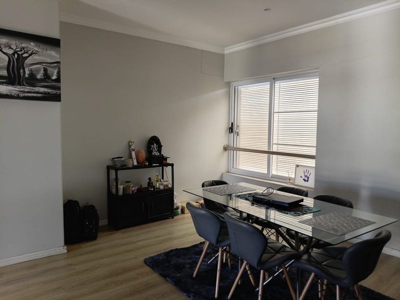 A dining room featuring light-toned laminate flooring, neutral walls, and a sliding window with security bars, providing a functional and well-lit space.
