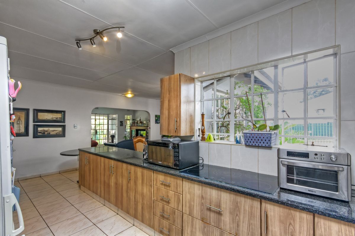 A functional kitchen featuring wood-look cabinetry, granite countertops, an electric hob, and tiled flooring, with an open-plan layout leading to the living area.
