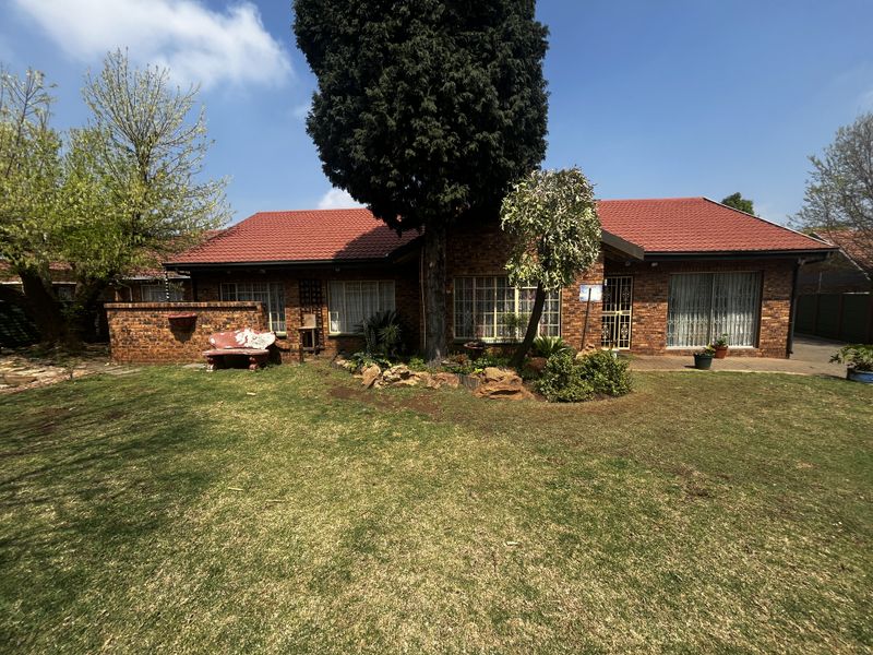 A rear view of a single-story brick house featuring a tiled roof, paved patio area, and a well-maintained lawn.