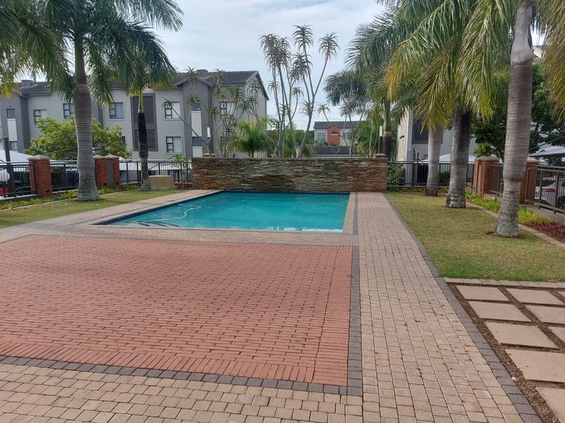 A well-maintained communal swimming pool area featuring a stone-clad water feature, paved decking, and established palm trees within a secure apartment complex.