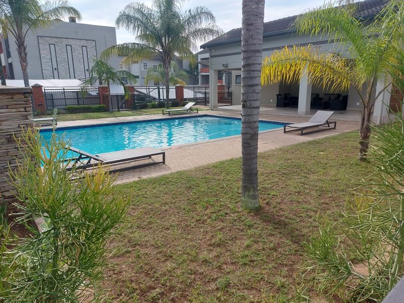 A well-maintained outdoor pool area featuring a paved deck, a covered patio space, and surrounding landscaped garden within a secure residential complex.