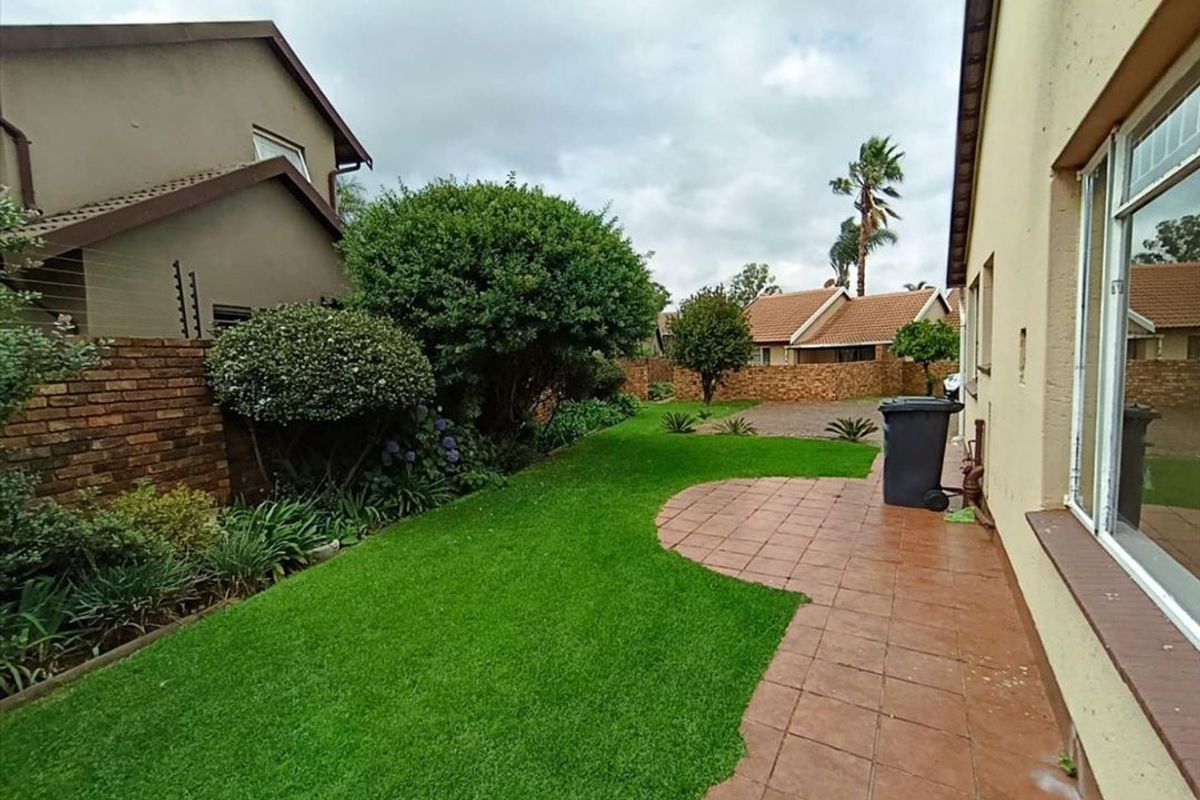 A rear exterior view of the townhouse featuring a paved patio area, a well-maintained lawn, and established garden beds enclosed by brick boundary walls.
