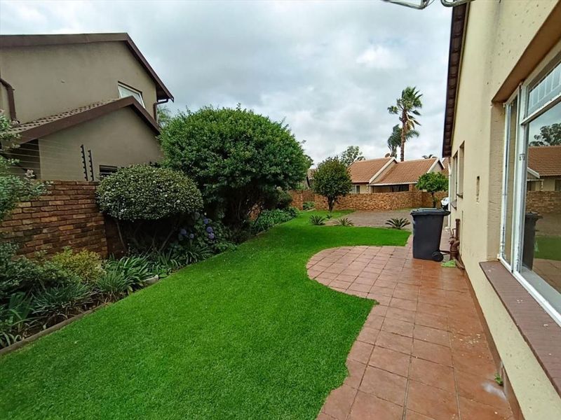 A rear exterior view of the townhouse featuring a paved patio area, a well-maintained lawn, and established garden beds enclosed by brick boundary walls.